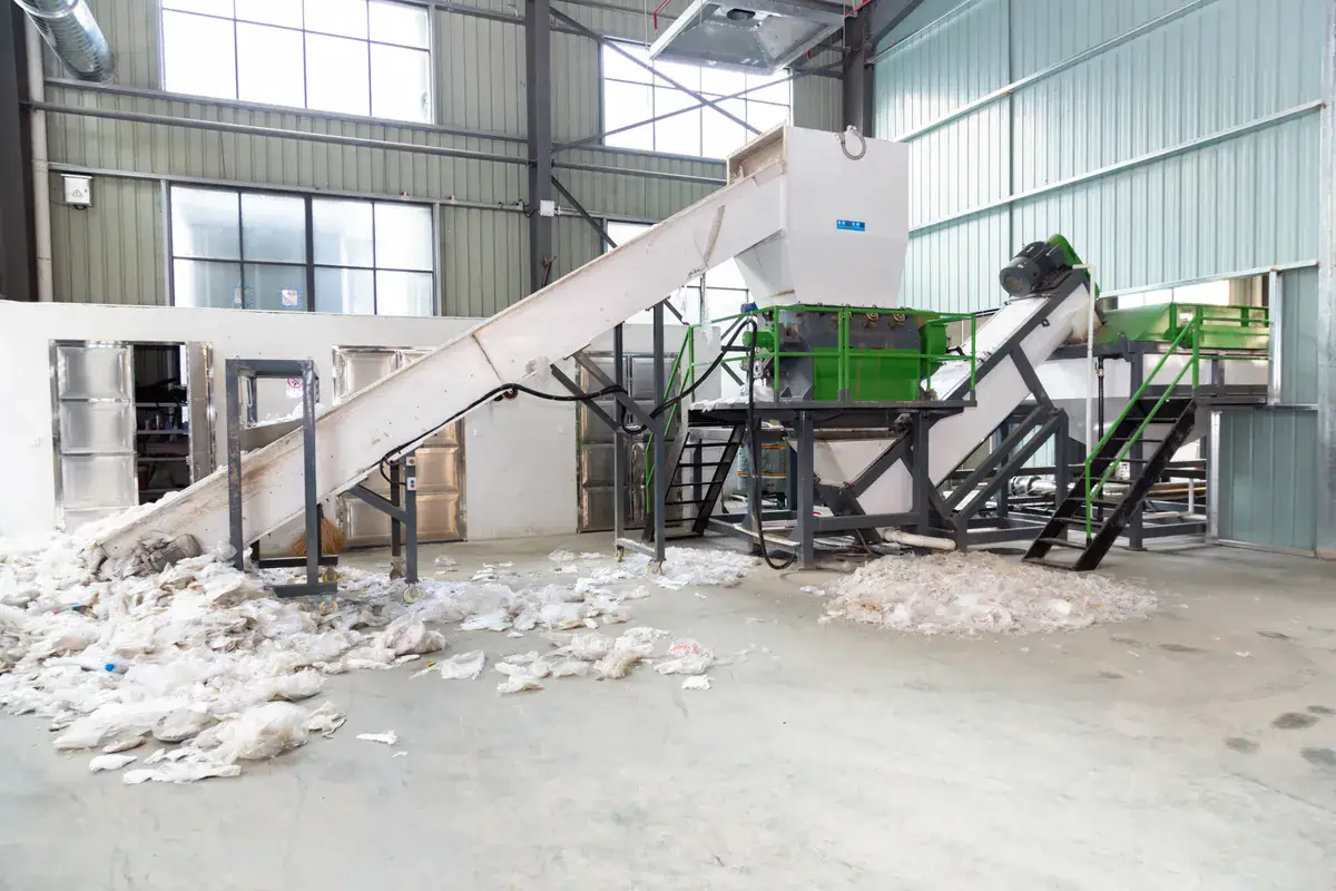 A shredding section in one of our film lines: the film is opened into pieces that move freely through the washers. Film washing line shredding section with a single-shaft shredder, conveyor and shredded film on the floor