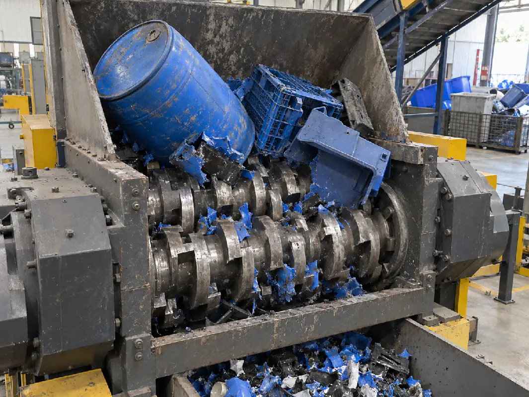 Double-shaft shredders pull bulky, irregular items such as drums and crates into the cutting zone from both sides. Double-shaft shredder grabbing blue plastic drums and crates in the hopper