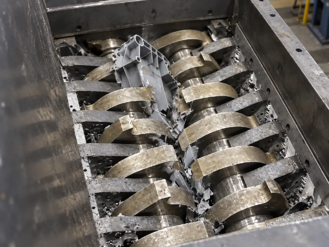 Double-shaft shredder cutters and hooks grabbing and tearing rigid plastic between two shafts. Two intermeshing shafts of a double-shaft shredder tearing a rigid plastic part